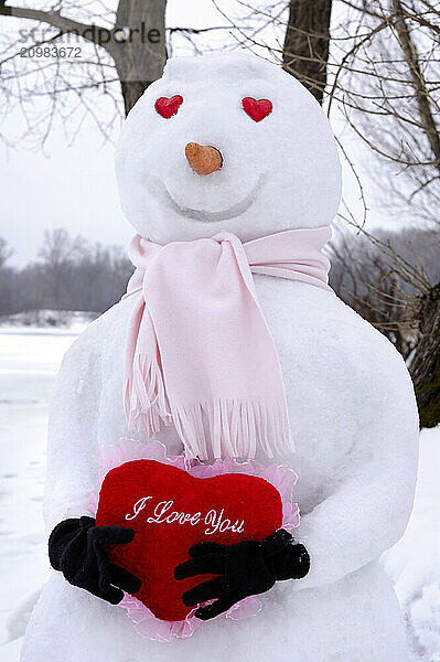 Smiling snowman with heart-shaped eyes holding a red heart with I Love You written on it in snow covered winter scenery. Valentines Day Romantic concept