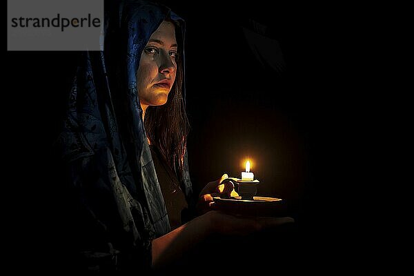 Sad young woman with candle and blue headscarf against a black background