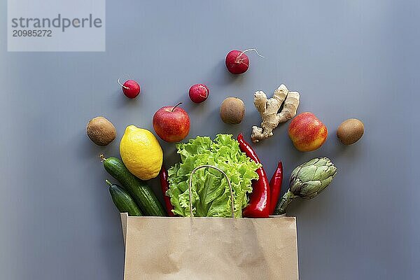 Healthy and organic food flay lay concept on gray background. Eco bag with scattered lettuce salad leaves  apples  kiwi  radish  lemon  cucumber  artichoke  red sweet pepper and ginger root