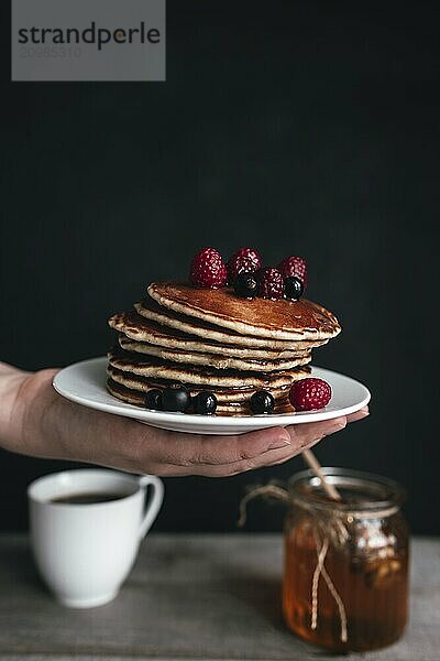 Juicy pancakes with berries and honey on white plate on human hand  jar and spoon  wooden table with cup of coffee.