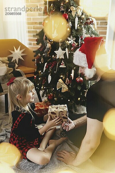 Young father in red Santa hat giving Christmas gift to his smiling daughter near decorated Christmas tree. Girl dressed in festive red-black Christmas outfit. They smiling  happy because of New Year