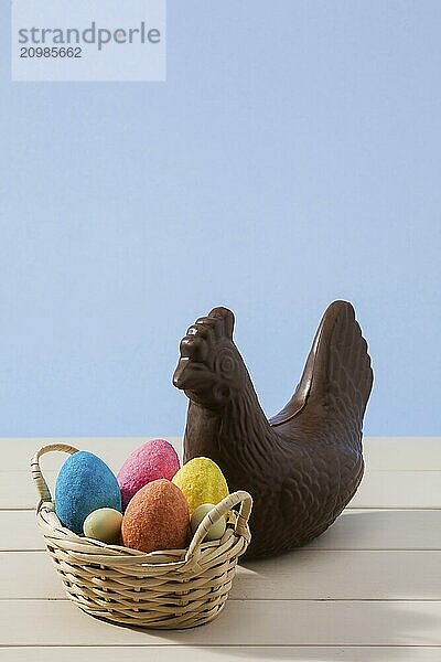 Easter chocolate chicken and colored eggs in a small basket over a white table with blue background