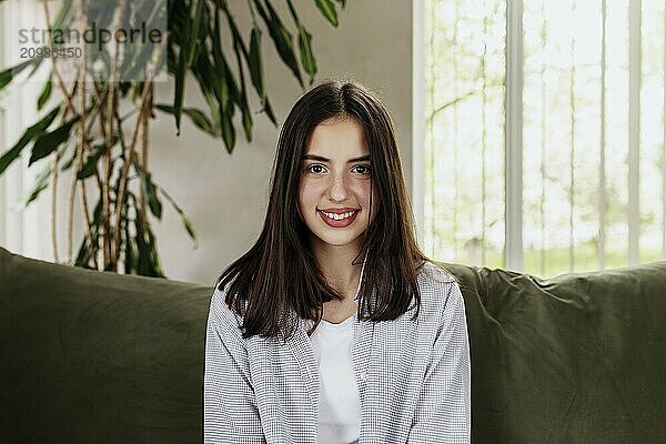 Beautiful smiling caucasian girl sitting at green sofa and watching at camera. Cute woman front view. Female smile  looking in photo camera. Online interview or meeting. Big lips.