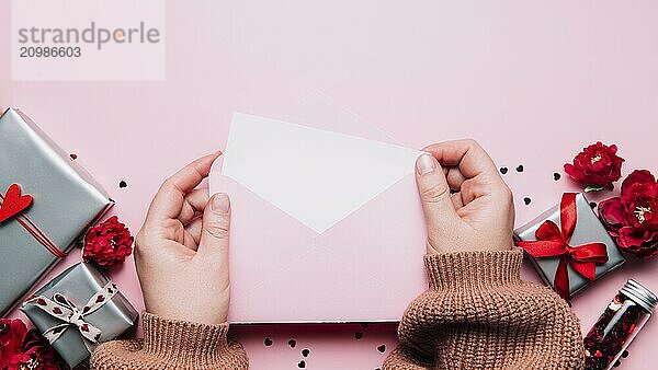 Female hands hold love letter empty message in open envelope  Valentine's Day greeting card or invitation blank mock up. Copy space  top view. Pink background with hearts  gray gift boxes  confetti