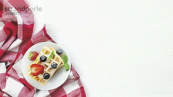 White plate on red kitchen towel with stack homemade Belgian waffles  blueberry  strawberry cuts  mint leaf  sugar powder on gray background