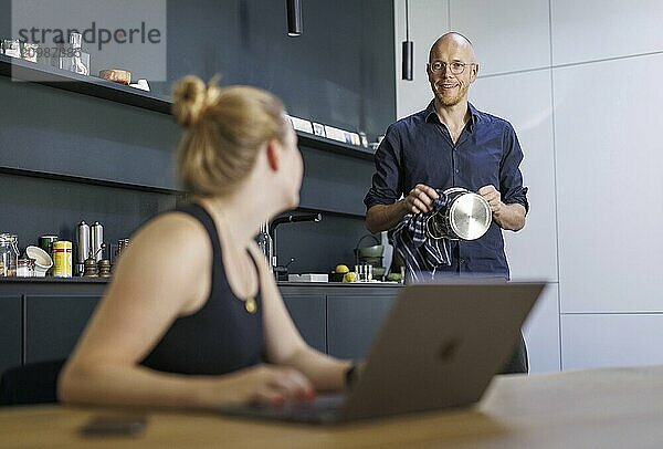 Symbolic photo on the subject of division of labour for couples in the household. A woman sits at a laptop in a kitchen while a man washes dishes in the background. Berlin  13.08.2024