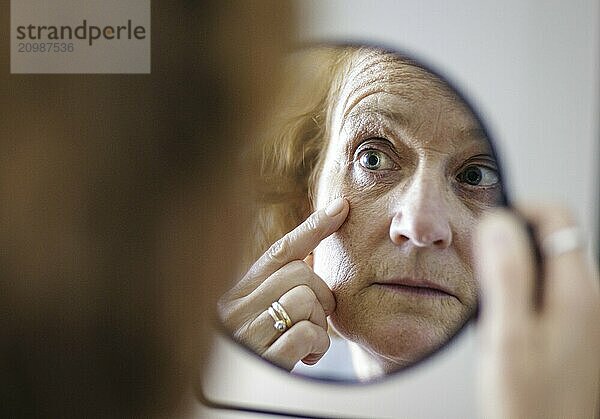 Symbolic photo on the subject of eye wrinkles. A woman looks at her eye wrinkles in the mirror. Berlin  13.08.2024