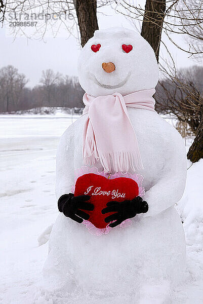 Smiling snowman with heart-shaped eyes holding a red heart with I Love You written on it in snow covered winter scenery