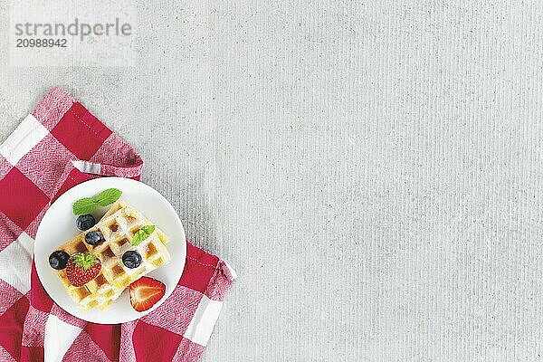 White plate on red kitchen towel with stack homemade Belgian waffles  blueberry  strawberry cuts  mint leaf  sugar powder on gray background