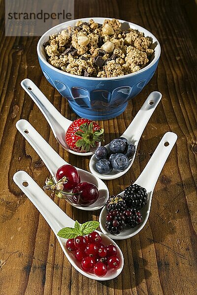 Collection of wild berries on white spoons and a cup of cereals over a wooden background