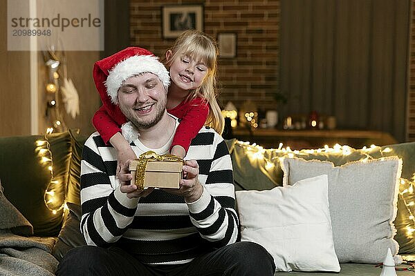 Merry Christmas and Happy New Year. Christmas gift surprise. A little smiling daughter girl hugging father back holding gift indoors at home decorated for holiday. Handsome man is holding giftbox