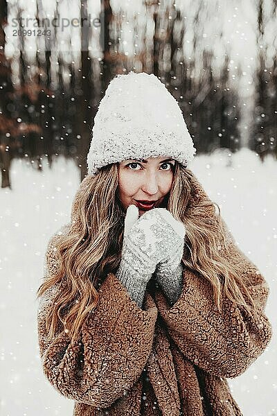 Beautiful young woman in snowy fancy winter woodland. Girl wearing fluffy gloves  cap and coat. Christmas forest  trees on blurred background. Crossed hands because of cold weather.