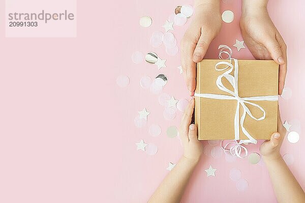 Flat lay with isolated hands giving a craft paper gift box tied with ribbon  star and circle paper confetti or glitters on pink pastel background. Adult and child holding giftbox.