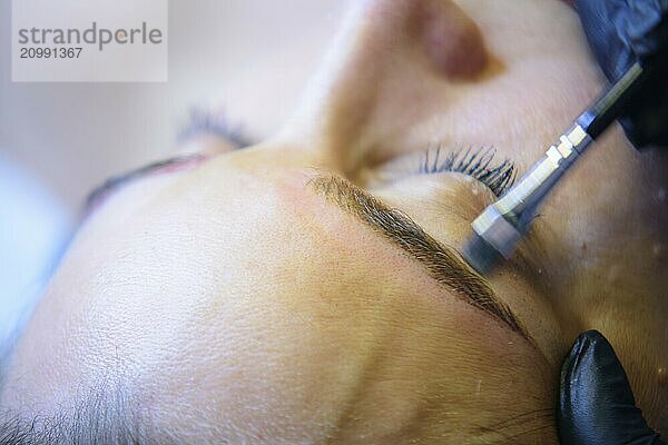 Beautician carefully applies permanent makeup to enhance a woman's eyebrows in a cozy salon. The close up image shows precise detail and promotes confidence