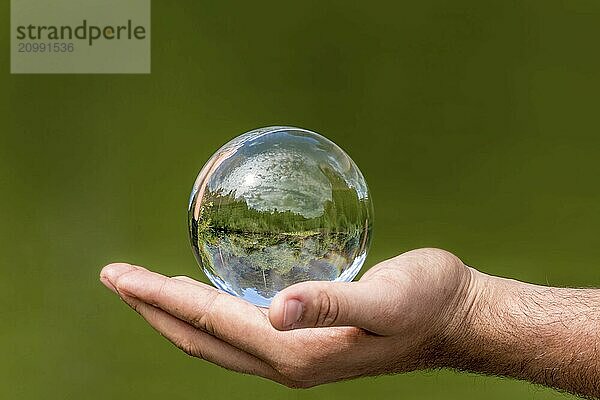 Glass ball with mirrored lake  trees and cloudy sky  lying in one hand against a green background