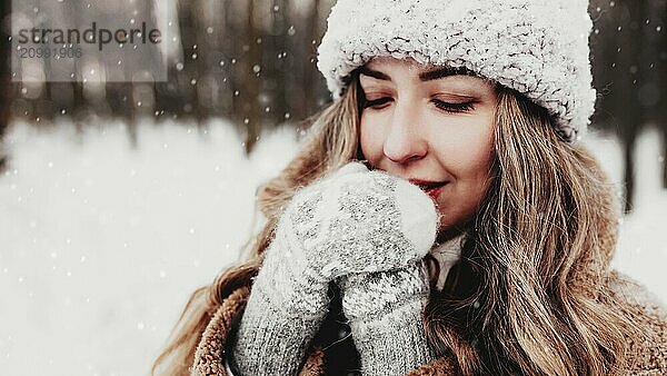 Beautiful young woman in snowy fancy winter woodland. Girl wearing fluffy gloves  cap and coat. Christmas forest  trees on blurred background. Crossed hands because of cold weather.