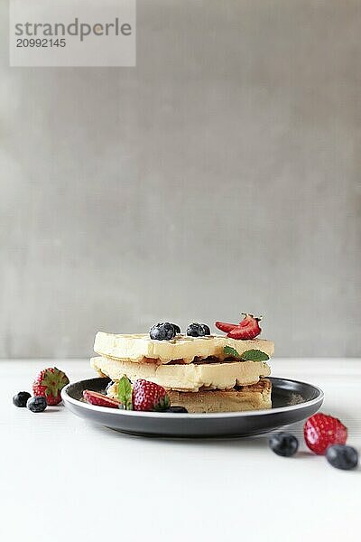 Stack of waffles on a plate on the white table with blueberry  chopped strawberry and mint leaves  gray background.