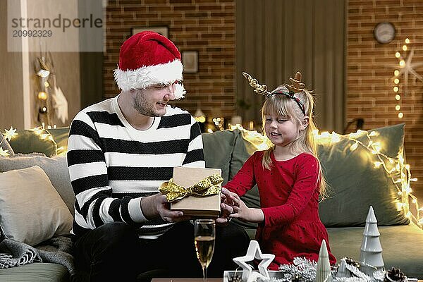 Merry Christmas and Happy New Year. Christmas gift surprise. A little happy smiling daughter girl opens a present. Father holding and giving giftbox indoors in house with festive Christmas decorations