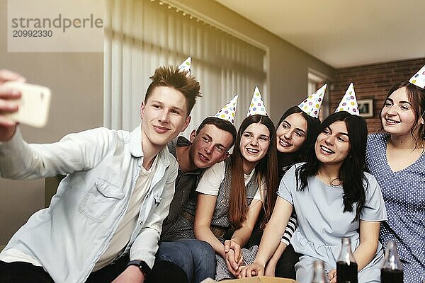 Group of best friends with festive cap taking selfie at birthday party. Girls and guys are posing and smiling at camera of mobile phone.