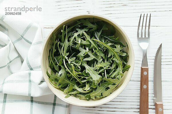 Green fresh arugula salad leaves in bowl  fork and knife on white wooden background  white towel. Healthy eating concept.