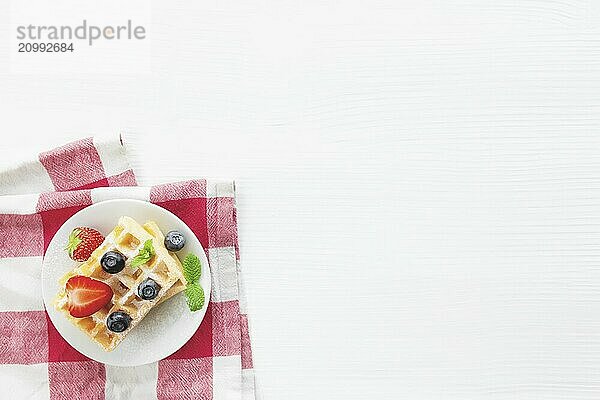 White plate on red kitchen towel with stack homemade Belgian waffles  blueberry  strawberry cuts  mint leaf  sugar powder on gray background