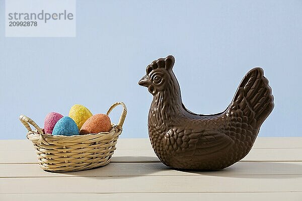 Easter chocolate chicken and colored eggs in a small basket over a white table with blue background