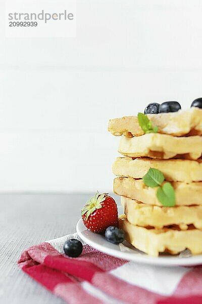 White plate on red kitchen towel with stack homemade Belgian waffles  blueberry  strawberry cuts  mint leaf  sugar powder on gray background