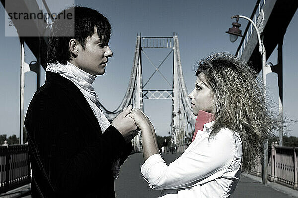 Young man holding hands of a young woman and looking at her eyes. Romantic young couple on the bridge