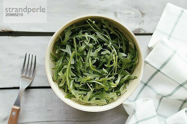 Green fresh arugula salad leaves in bowl  fork on white wooden background  white towel. Healthy eating concept.