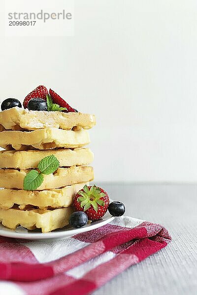 White plate on red kitchen towel with stack homemade Belgian waffles  blueberry  strawberry cuts  mint leaf  sugar powder on gray background