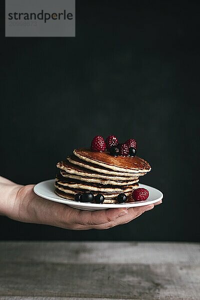 Juicy pancakes with berries and honey on white plate on human hand  wooden table.