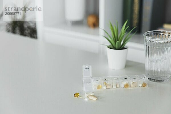 Closeup of medical pill box with doses of tablets for daily take a medicine for treatment  cure the disease. Glass cup of water on table  gray background. Green flowerpot or plant. Helathy lifestyle