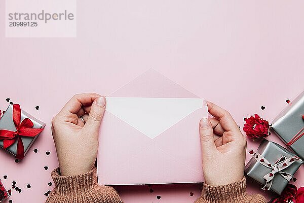 Female hands hold love letter empty message in open envelope  Valentine's Day greeting card or invitation blank mock up. Copy space  top view. Pink background with hearts  gray gift boxes  confetti