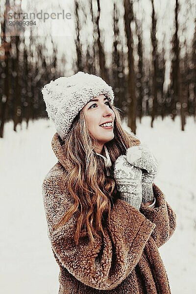 Beautiful young woman in snowy fancy winter woodland. Girl wearing fluffy gloves  cap and coat. Christmas forest  trees on blurred background. Crossed hands because of cold weather.