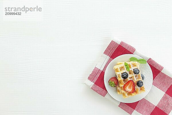 White plate on red kitchen towel with stack homemade Belgian waffles  blueberry  strawberry cuts  mint leaf  sugar powder on gray background