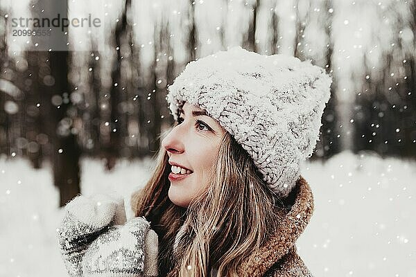 Beautiful young woman in snowy fancy winter woodland. Girl wearing fluffy gloves  cap and coat. Christmas forest  trees on blurred background. Crossed hands because of cold weather.