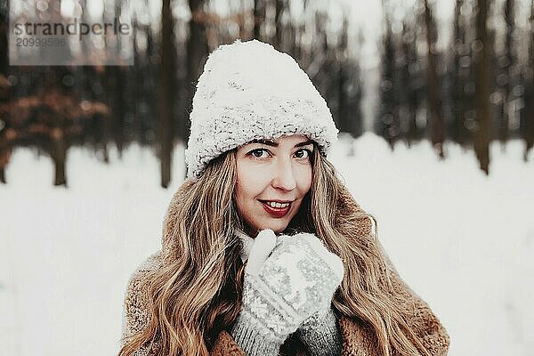 Beautiful young woman in snowy fancy winter woodland. Girl wearing fluffy gloves  cap and coat. Christmas forest  trees on blurred background. Crossed hands because of cold weather.