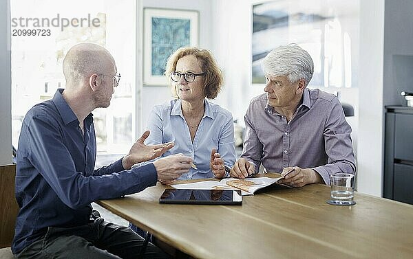 Symbolic photo on the subject of counselling. An older woman and an older man sit together at a table at home and receive counselling from a young man. Berlin  13.08.2024