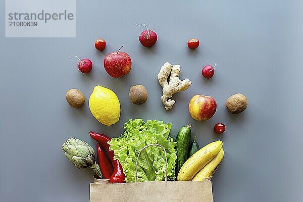 Healthy and organic food flay lay concept on gray background. Eco bag with scattered lettuce salad leaves  apples  kiwi  radish  lemon  cucumber  banana  artichoke  red sweet pepper and ginger root