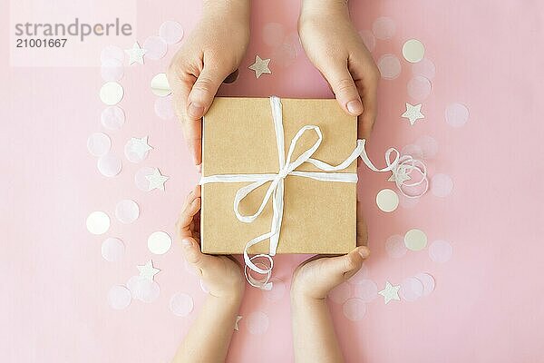 Flat lay with isolated hands giving a craft paper gift box tied with ribbon  star and circle paper confetti or glitters on pink pastel background. Adult and child holding giftbox.