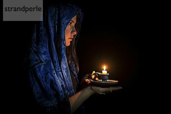 Sad young woman with candle and blue headscarf against a black background