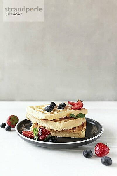 Stack of waffles on a plate on the white table with blueberry  chopped strawberry and mint leaves  gray background.