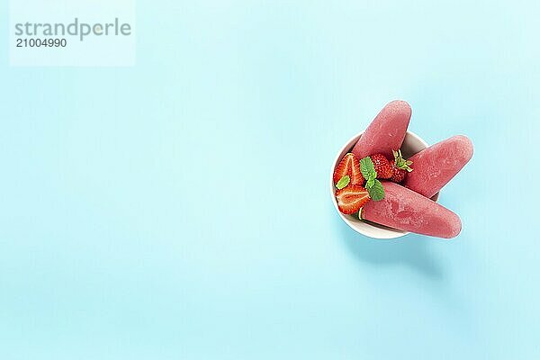 Top view on bowl with fruity pink frozen sorbet ice-cream on plastic stick with strawberry  mint on blue background. Frozen mixed fruits with yogurt in a form of ice-cream. Kids love to eat it a lot