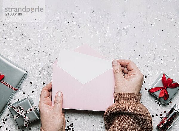 Female hands hold love letter empty message in open envelope  Valentine's Day greeting card or invitation blank mock up. Copy space  top view. Pink background with hearts  gray gift boxes  confetti