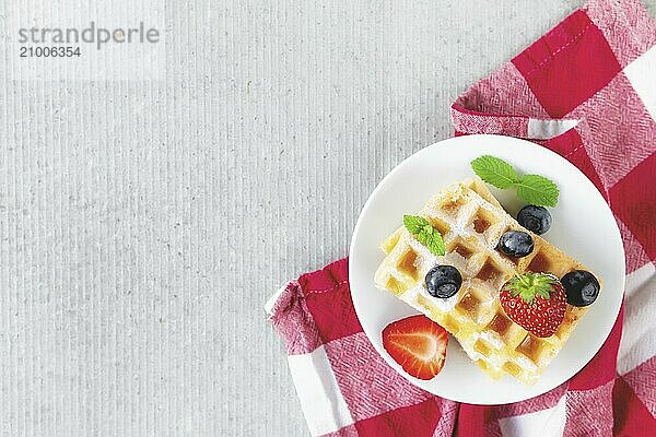 White plate on red kitchen towel with stack homemade Belgian waffles  blueberry  strawberry cuts  mint leaf  sugar powder on gray background