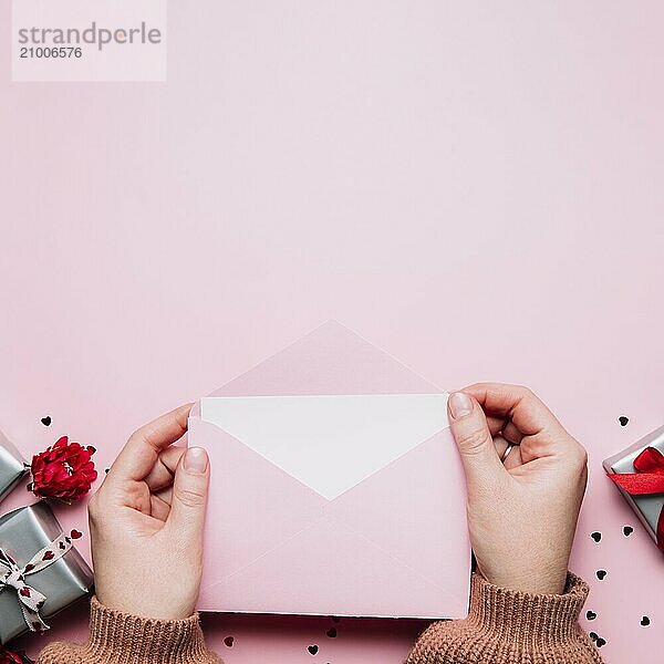 Female hands hold love letter empty message in open envelope  Valentine's Day greeting card or invitation blank mock up. Copy space  top view. Pink background with hearts  gray gift boxes  confetti