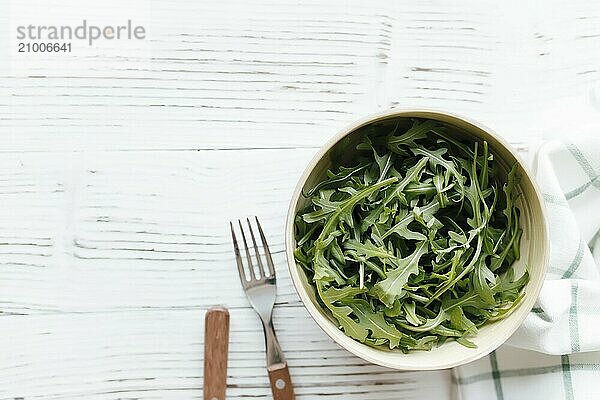 Green fresh arugula salad leaves in bowl  fork and knife on white wooden background  white towel. Healthy eating concept.