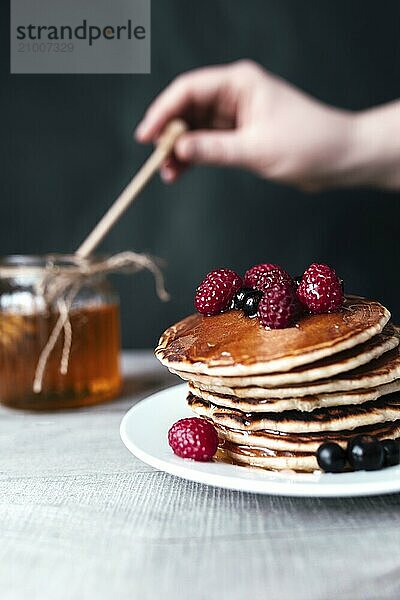 Pancakes with berries and honey on white plate  hand holding spoon in jar  wooden table.