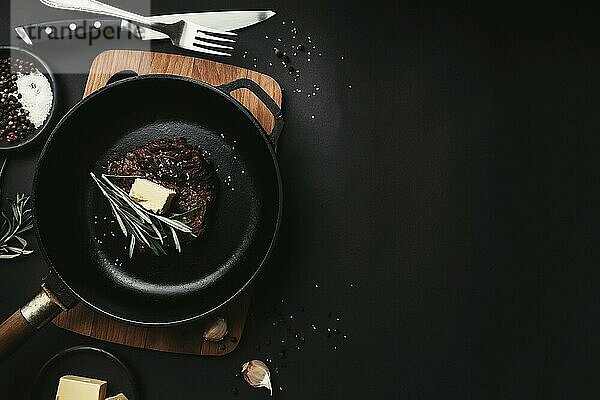 Cooked ribeye steak on cast-iron pan and board with pepper  rosemary  salt  garlic  knife  fork  butter on black background