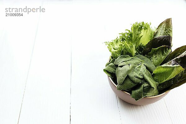 Flat lay with bowl of fresh green salad leaves of spinach and lettuce  romaine and basil on white wooden background. Healthy vegetarian eating concept.